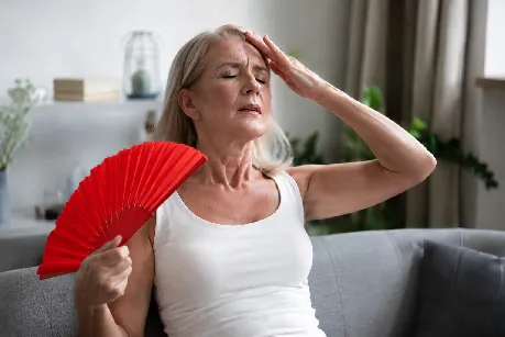 Vrouw van middelbare leeftijd zit op de bank en wuift zichzelf koelte toe met een rode waaier, terwijl zij haar hand tegen haar voorhoofd houdt.