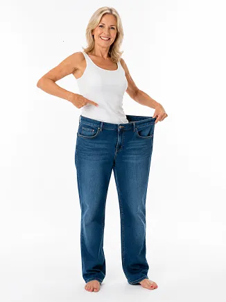 Vrouw in wit mouwloos shirt en blauwe jeans, wijzend naar de broek om de pasvorm te tonen